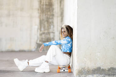 Ganzkörper-Seitenansicht von stilvollen jungen Hipster afroamerikanische Frau in trendigen Klamotten und Stiefel sitzen auf Skateboard gegen Betonwand, während auf städtischen Straße ausruhen - ADSF22913