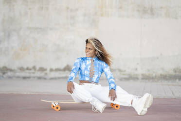 Full body stylish young hipster African American female in trendy clothes and boots sitting on skateboard against concrete wall while resting on urban street - ADSF22916