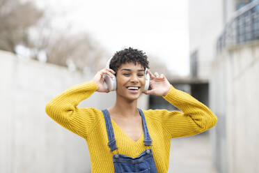 Verträumt lächelnde Afroamerikanerin mit kabellosen Kopfhörern, die auf der Straße stehend Lieder hört - ADSF22919