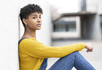 Seitenansicht einer positiven Afroamerikanerin in Jeans-Overall und mit Afrofrisur, die sich an eine Wand lehnt und wegschaut - ADSF22922