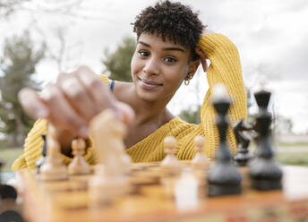 Von unten optimistische afroamerikanische Frau, die an einem Tisch im Park sitzt und Schach spielt, während sie in die Kamera schaut - ADSF22924