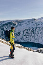 Full body side view of male mountaineer in warm activewear with backpack standing on slope of snowy rocky mountain and enjoying spectacular landscape in sunny winter day - ADSF22932