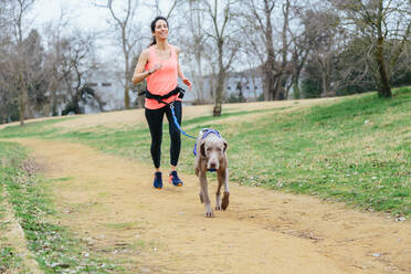 Full body of positive young fit female running together with Weimaraner dog during outdoor workout in park - ADSF22934