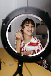Smiling female influencer applying make-up while vlogging at studio - TCEF01763