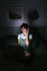 Lächelnde Frau mit Kopfhörern und Laptop in der Dunkelkammer - TCEF01772