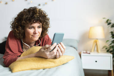 Frau, die ein Mobiltelefon benutzt, während sie im Schlafzimmer zu Hause über Kopfhörer Musik hört - JPTF00765