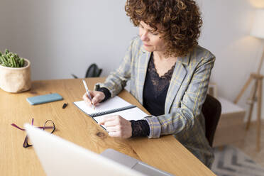 Curly haired woman writing in book on table at home - JPTF00776