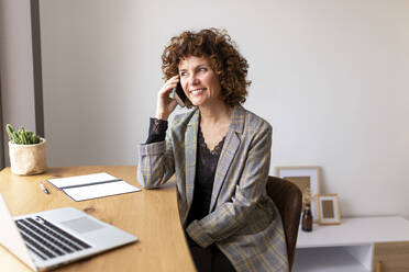 Smiling female freelancer talking on smart phone while sitting on chair at home - JPTF00780