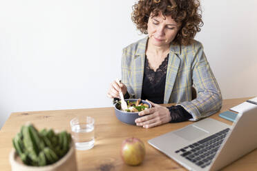 Mature female freelancer sitting at table with laptop having food at home - JPTF00782