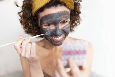 Lächelnde reife Frau beim Auftragen der Gesichtsmaske im Schlafzimmer zu Hause - JPTF00802