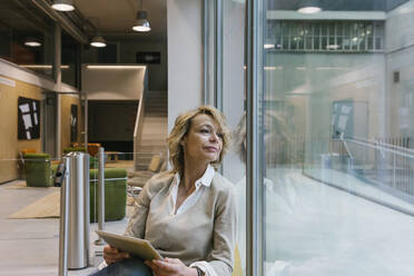 Berufstätige Frau träumt am Fenster sitzend mit digitalem Tablet in einem Coworking-Büro - XLGF01465