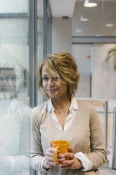 Mature businesswoman with blond hair having coffee while standing by window in office - XLGF01466