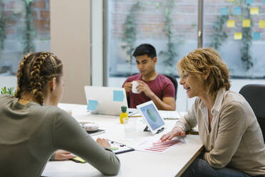 Female professionals discussing over business plan while businessman having coffee at desk - XLGF01516