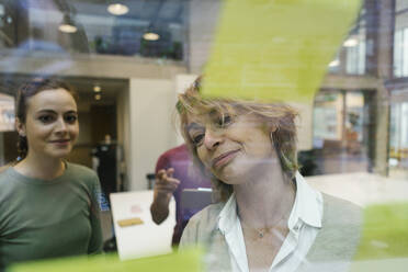 Businesswomen looking at adhesive notes on glass at office seen through glass - XLGF01521