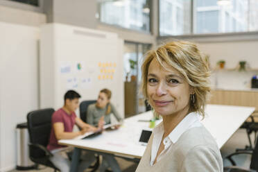Geschäftsfrau lächelnd, während männliche und weibliche Mitarbeiter im Hintergrund im Büro diskutieren - XLGF01526
