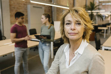 Reife Geschäftsfrau mit blondem Haar mit männlichen und weiblichen Mitarbeitern im Hintergrund im Büro - XLGF01534