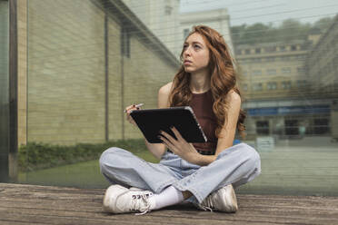Nachdenkliche Frau hält digitales Tablet, während sie vor einer Glaswand sitzt - MTBF00958