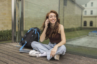 Lächelnde junge Frau, die mit dem Handy telefoniert, während sie an einem Rucksack sitzt - MTBF00961
