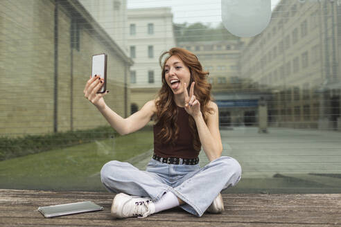 Frau gestikuliert bei der Aufnahme eines Selfies mit dem Handy vor einer Glaswand - MTBF00964