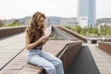 Young woman using mobile phone while sitting on retaining wall - MTBF00967