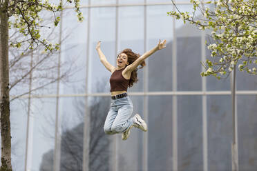 Carefree woman jumping with arms outstretched in front of glass building - MTBF00975