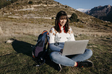 Aufgeregte Frau, die einen Laptop benutzt, während sie im Gras auf einem Berg sitzt - TCEF01783