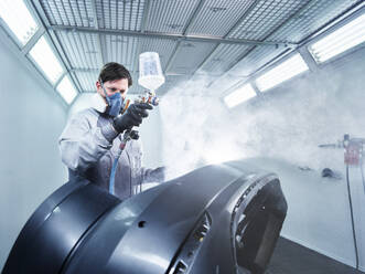 Männlicher Spritzlackierer mit Gesichtsschutzmaske bei der Arbeit mit Airbrush an einem Fahrzeugteil in einer Werkstatt - CVF01729
