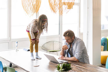 Businessman using laptop by daughter standing on table at home - JOSEF04130