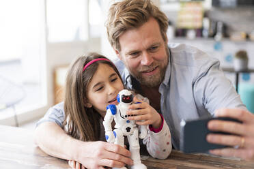 Father taking selfie through mobile phone with daughter and robot toy at home - JOSEF04134