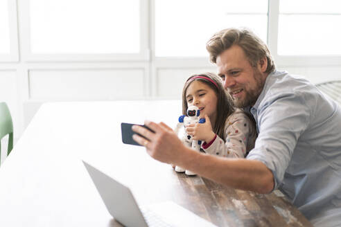 Vater, der ein Selfie mit seiner Tochter macht, die einen Spielzeugroboter über ein Mobiltelefon zu Hause hält - JOSEF04158