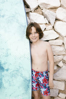 Smiling boy standing with surfboard during summer - AGGF00090