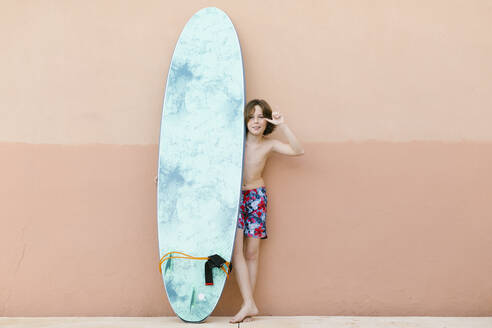 Boy wearing swimming trunks gesturing while standing with surfboard in front of beige wall - AGGF00092