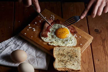 From above of crop anonymous person chopping roast eggs and bacon with knife and fork for breakfast - ADSF22961