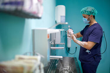 Side view of anonymous young male doctor in medical uniform and mask washing hands with soap before surgery in operating room - ADSF22964