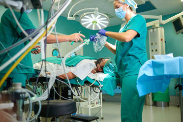 Side view of crop anonymous female assistants in medical uniforms and masks preparing for giving anesthesia to patient lying on couch in operating room - ADSF22967