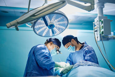 Seitenansicht eines nicht erkennbaren männlichen Arztes mit Assistenten in medizinischen Kitteln und Masken bei der Durchführung einer Operation mit Laser im Operationssaal - ADSF22970
