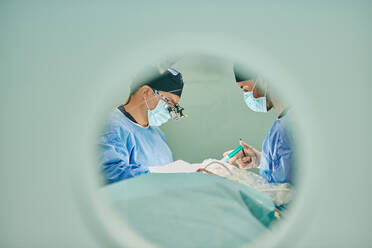 Durch das runde Fenster von unkenntlichen Ärzten in Uniformen und Masken, die in einem modernen Operationssaal operieren - ADSF22979
