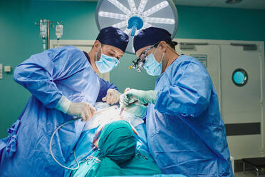 Side view of unrecognizable young concentrated male assistant in sterile mask and gown giving instruments to plastic surgeon performing rhinoplasty in operating room - ADSF22980