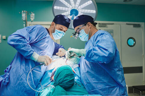 Seitenansicht eines nicht erkennbaren jungen, konzentrierten männlichen Assistenten mit steriler Maske und Kittel, der dem plastischen Chirurgen, der eine Nasenkorrektur vornimmt, im Operationssaal Instrumente reicht - ADSF22980