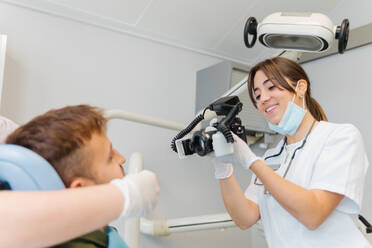 Smiling competent female dentist taking photos of male patient teeth while working in contemporary equipped dental clinic - ADSF22986