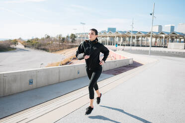 Sportliche Ganzkörper-Joggerin in schwarzer Sportkleidung, die auf einer asphaltierten Straße in einem sonnigen Vorort läuft und wegschaut - ADSF22987