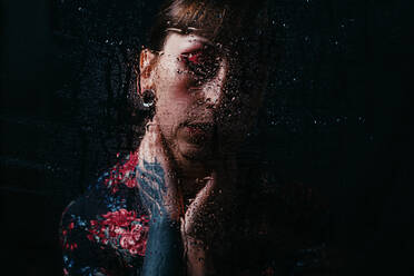 Aufgewühlte Frau mit geschlossenen Augen und gemaltem Arm, der den Hals berührt, hinter durchsichtigem Glas mit Wassertropfen - ADSF22998