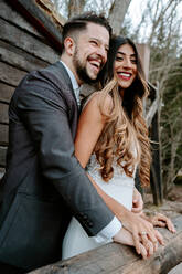 Side view of cheerful young bearded groom in suit laughing and cuddling elegant ethnic bride in white dress while standing together on wooden bridge near aged house in autumn forest - ADSF23014