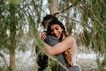 Side view of young ethnic bride with long hair in white dress smiling and hugging groom with closed eyes while standing together near coniferous tree in forest - ADSF23016