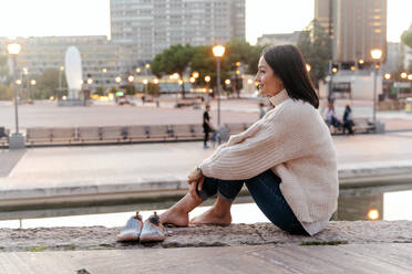 Side view of tender tranquil female embracing knees while sitting on stone embankment in city in evening and looking away - ADSF23017