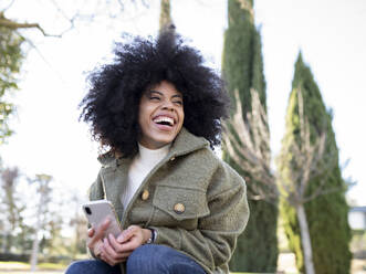 Low angle of charismatic young African American female millennial with curly hair laughing and looking away while using smartphone in park on sunny day - ADSF23029