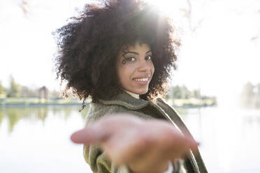 Seitenansicht einer positiven jungen schwarzen Frau mit Afrofrisur in trendigem Outfit, die ihre Hand in Richtung Kamera ausstreckt und lächelt, während sie sich in der Nähe eines Sees im Herbstpark ausruht - ADSF23031