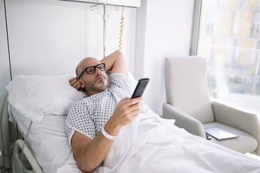 Erwachsener männlicher Patient im Krankenhauskittel auf dem Bett liegend und mit Fernbedienung in einer hellen Station in einer Klinik - ADSF23039