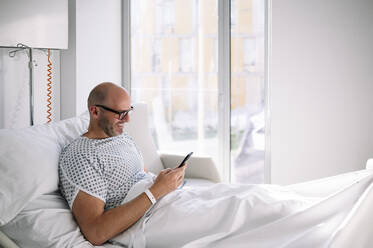 Positiver erwachsener Mann mit Patientenkittel und Brille, der auf einem Bett in einer hellen Station in einem modernen Krankenhaus telefoniert - ADSF23040