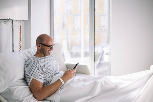 Positive adult male wearing patient gown and eyeglasses browsing phone on bed in light ward in modern hospital - ADSF23040